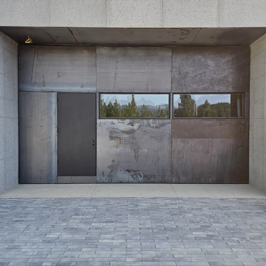 Modern concrete and metal structure with a dark door and reflective horizontal windows showing trees and mountains.