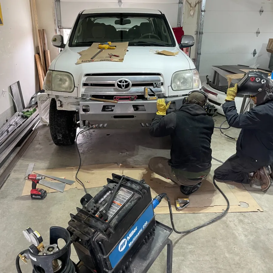 Two mechanics welding the front bumper of a white Toyota SUV inside a garage workshop.