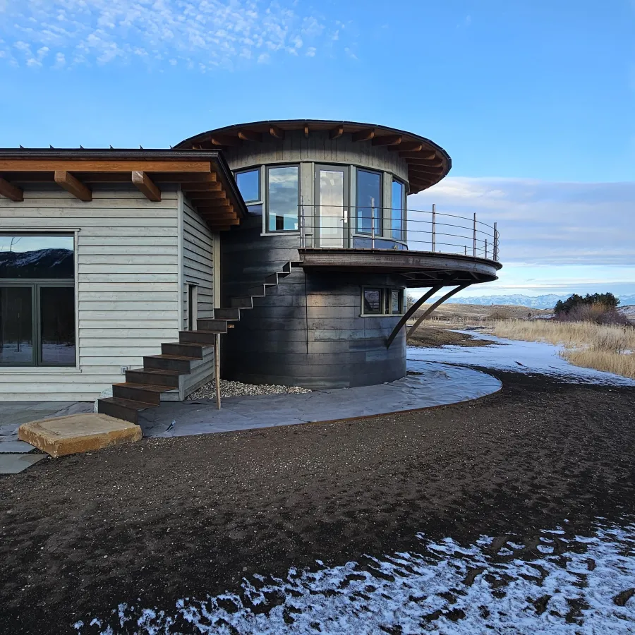 Modern round black metal building with wraparound balcony next to wood-paneled house under blue sky and light snow.