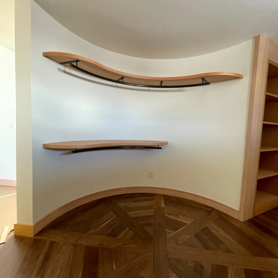 Curved wooden shelves with black brackets mounted on a white wall above polished wood parquet flooring.