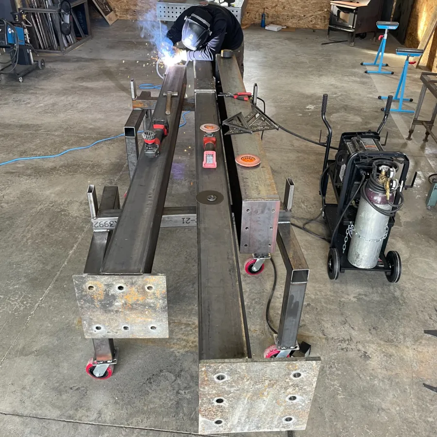 Welder working on large steel beams in a workshop with welding equipment and concrete floor.