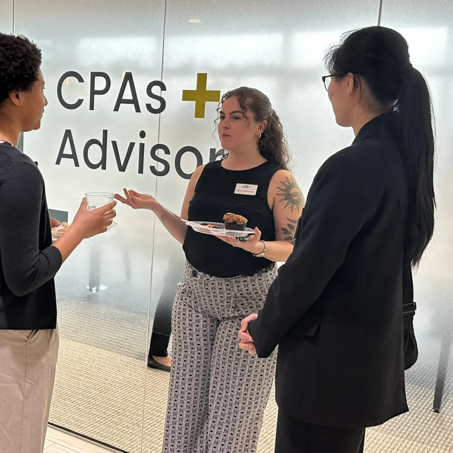 Three professional women networking and conversing in front of a glass wall with CPAs and Advisors signage.