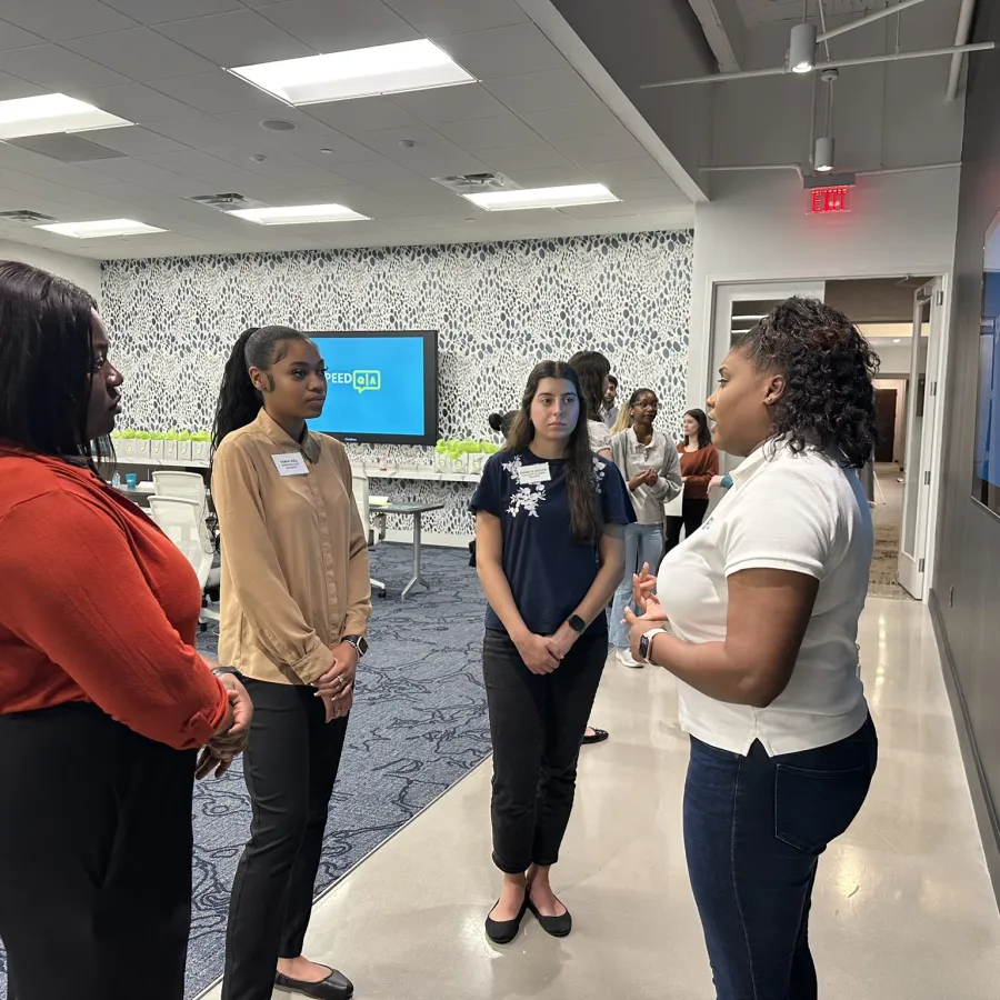 A woman in white shirt talks to three women standing and listening in a modern office space with monitors and chairs.