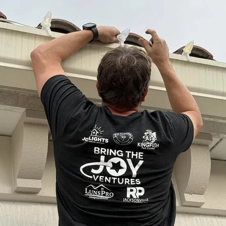 Man wearing a Bring The Joy Ventures t-shirt installs string lights on a house gutter under a tiled roof.