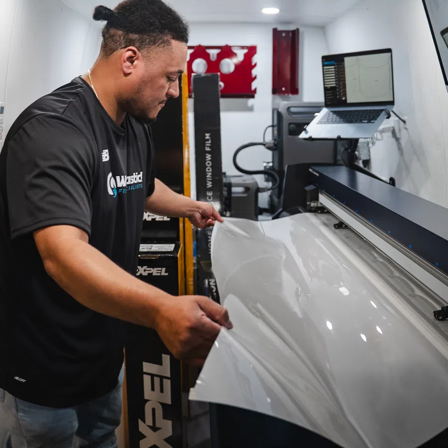 Technician handling clear protective film near cutting machine with laptop in a workshop environment