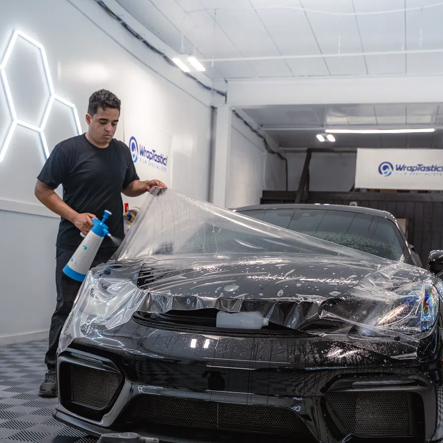 Man applying clear protective film on black sports car hood in a car detailing shop.