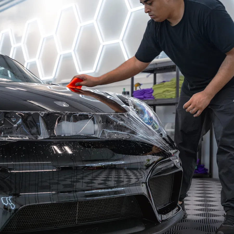 Man applying protective film on the hood of a black sports car in a modern garage with hexagonal lights.