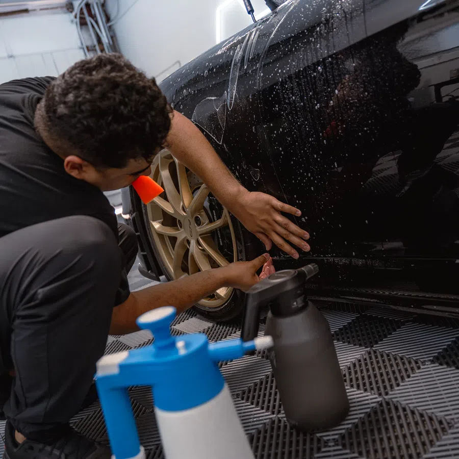 Man detailing black sports car with spray bottles on garage floor and water droplets on car surface