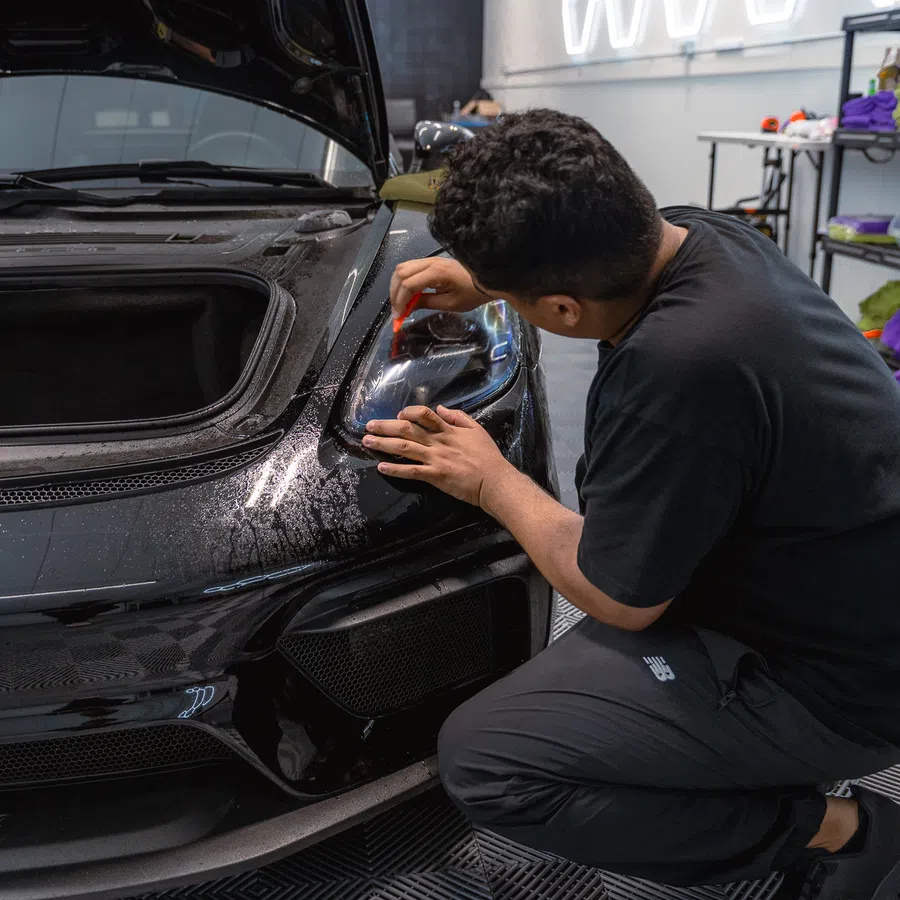 Man detailing a black sports car headlight in a modern garage with hexagonal light fixtures on the wall.