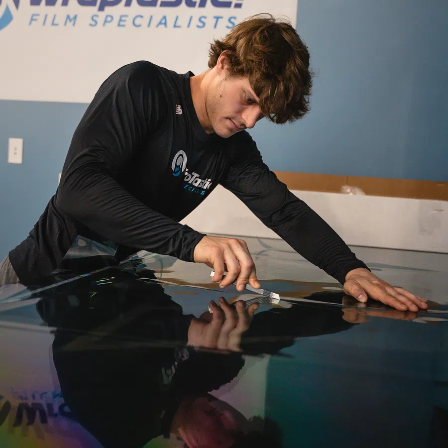 Man carefully applying window film on a glass surface in a WrapTastic workshop with logo on wall