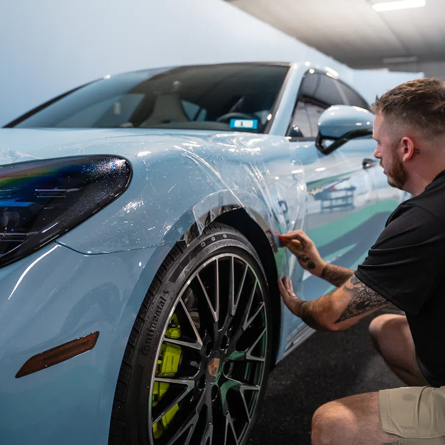 Man applying protective film to front side of a light blue sports car inside a garage.