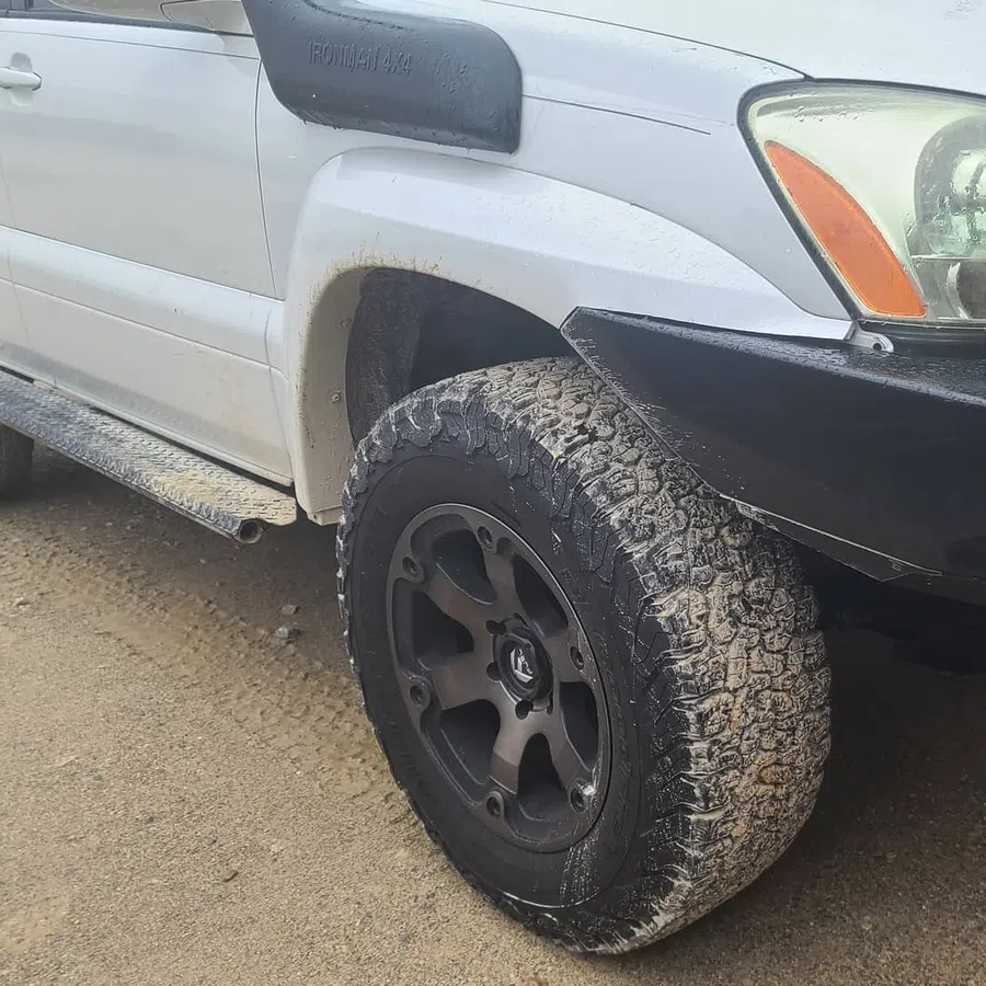 Close-up of a white off-road SUV front tire with mud and black custom rims on a dirt road.
