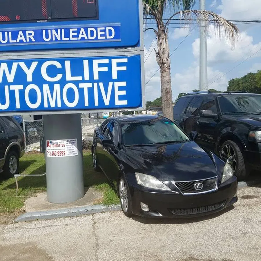 Black Lexus car parked in front of Wycliff Automotive sign with palm tree and blue sky background.