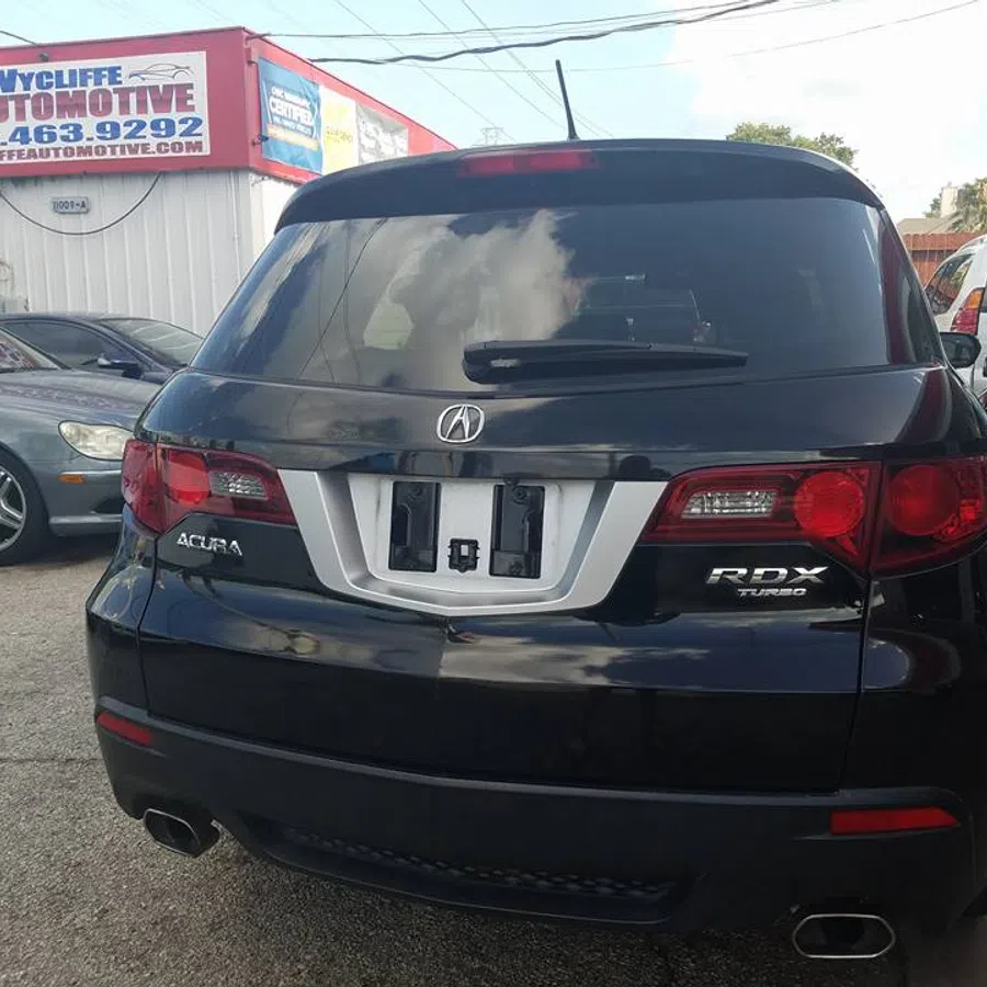 Rear view of a black Acura RDX Turbo SUV parked in a lot with other cars and automotive signage.