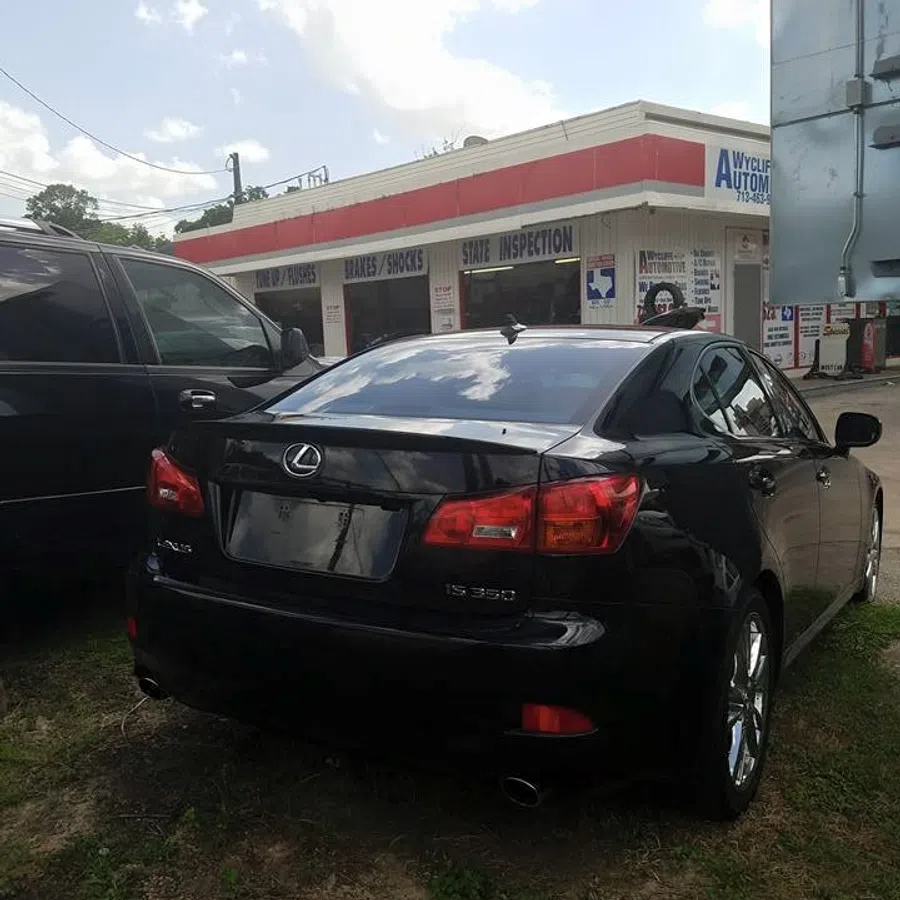 Black Lexus IS 350 parked near a repair shop with signs for brakes, shocks, and state inspection under a cloudy sky.