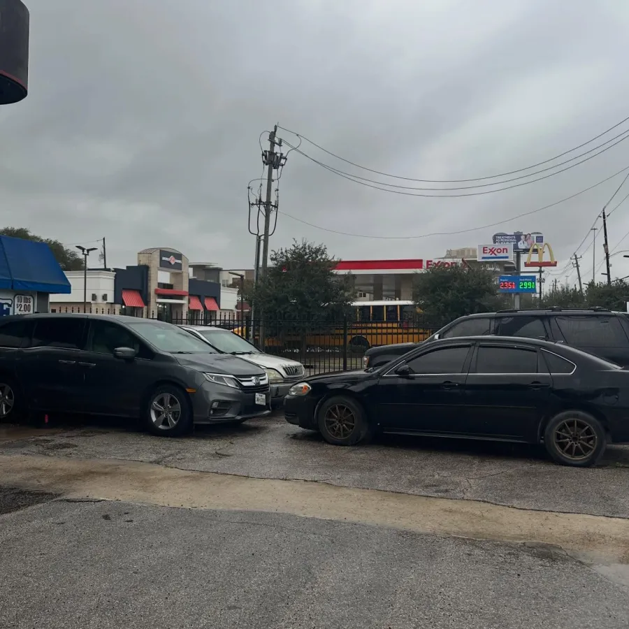 Wet parking lot with three cars, overhead power lines, a school bus, and an Exxon gas station with visible prices.