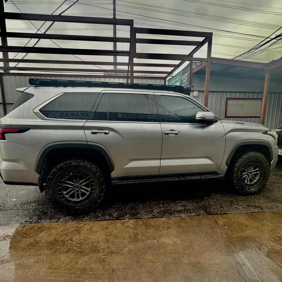 Silver off-road SUV parked on wet concrete under metal frame structure with cloudy sky.