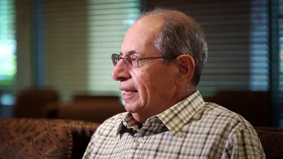 Elderly man with glasses wearing a checkered shirt, sitting indoors with soft lighting and blinds in the background.