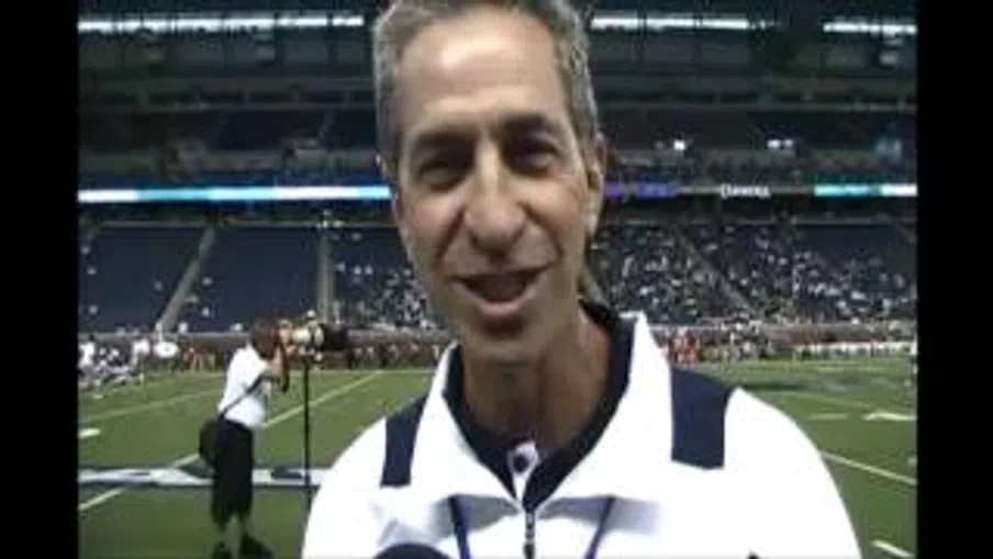 Man in white and navy sports jacket speaking on a football field with stadium and crowd in the background