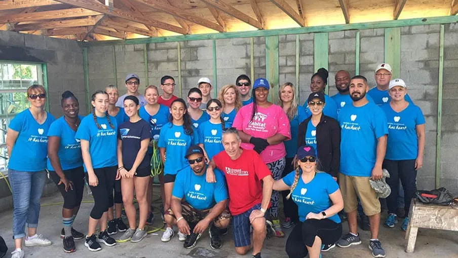 Group of diverse volunteers wearing blue and pink shirts posing inside an unfinished house under construction.