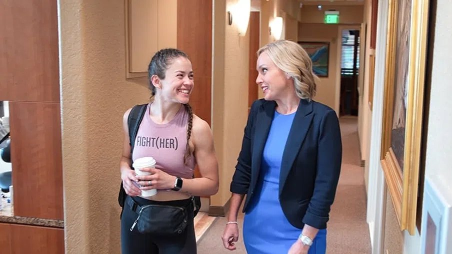 Two women smiling and walking together in an office hallway, one in athletic wear and the other in business attire.