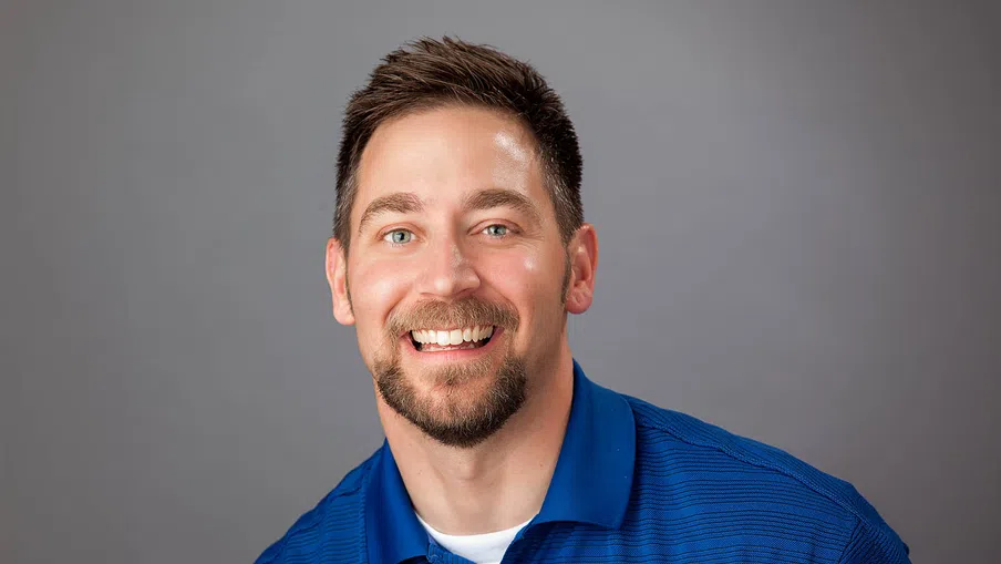Smiling man with short brown hair and beard wearing a blue polo shirt against a gray background