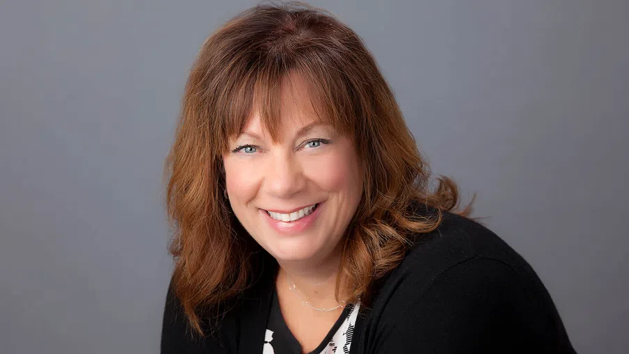 Smiling middle-aged woman with brown hair wearing a black cardigan and patterned blouse against a gray background