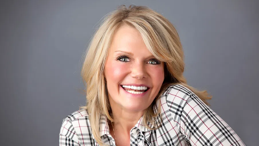 Smiling blonde woman wearing a plaid shirt against a plain gray background, looking directly at the camera.