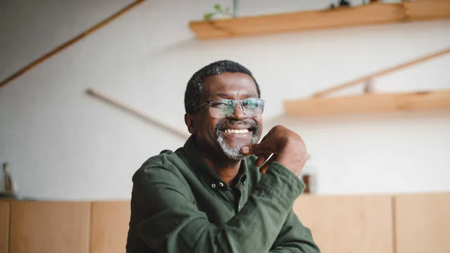Smiling middle-aged man with glasses and gray beard sitting indoors wearing a green shirt, relaxed and thoughtful.