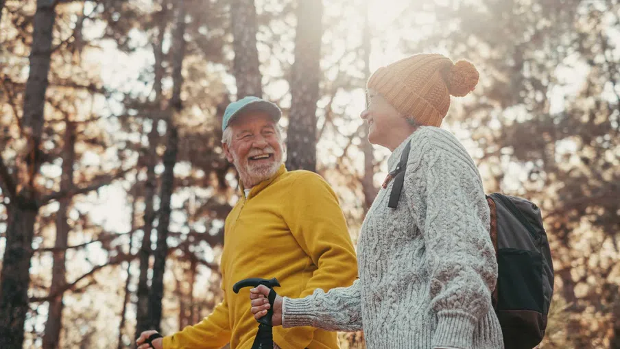 Senior couple hiking in autumn forest wearing warm clothes and carrying trekking poles, smiling and enjoying nature.