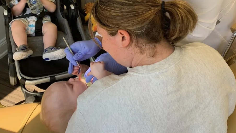 Dentist wearing mask and gloves examining a baby held by a woman while another baby sits in stroller.