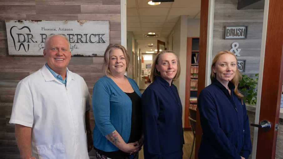 Team photo of four dental professionals standing inside a clinic with a wooden accent wall and sign