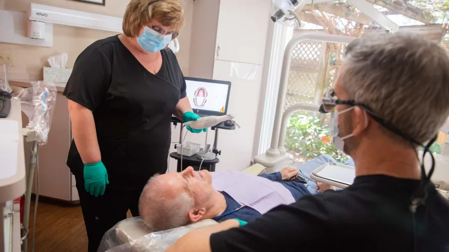 Dentist and assistant examining senior male patient in dental clinic with equipment and natural light.