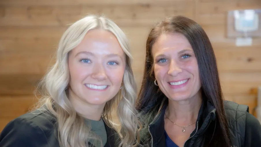 Two smiling women with blonde and brunette hair in casual jackets posing indoors with wooden background.