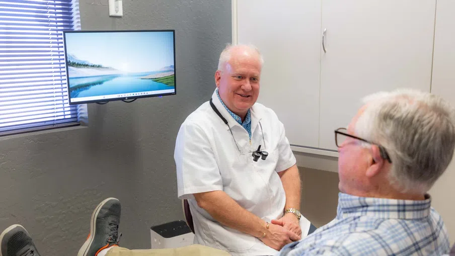 Dentist consulting an elderly male patient in a bright, modern dental office with a scenic window view on the monitor.