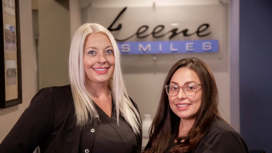 Two smiling dental professionals in black uniforms standing inside a dental clinic reception area.