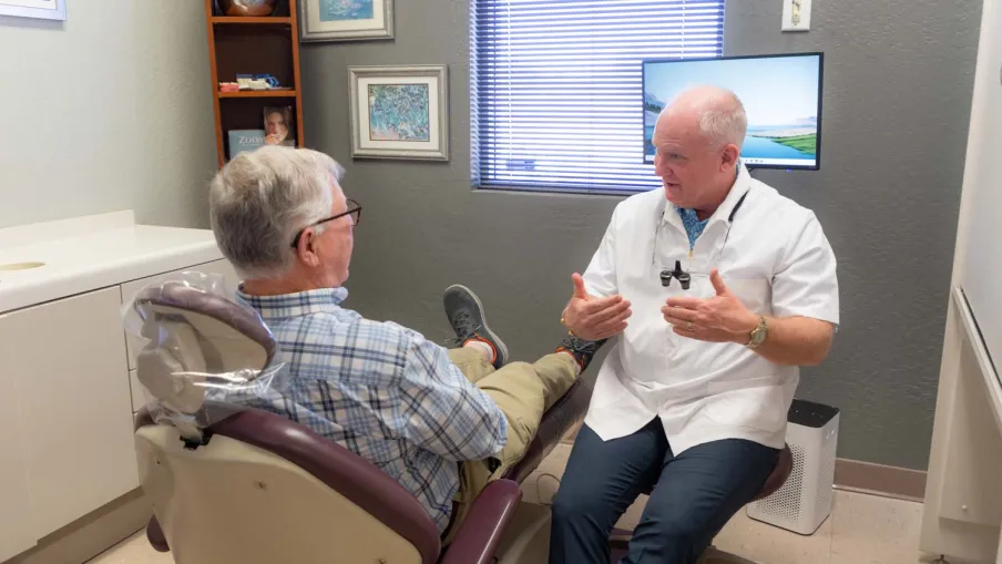 Dentist discussing treatment with elderly male patient in dental office consultation room.