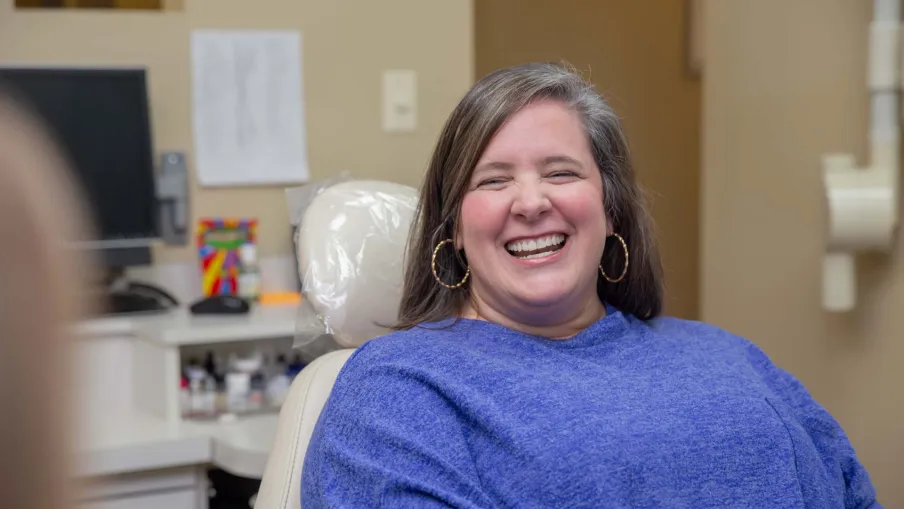 Smiling woman wearing a blue sweater sitting in a dental chair in a bright clinic environment