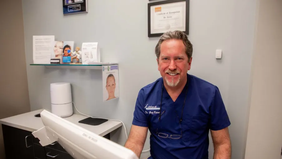 Smiling male doctor in blue scrubs sitting at desk with computer in a medical office setting.