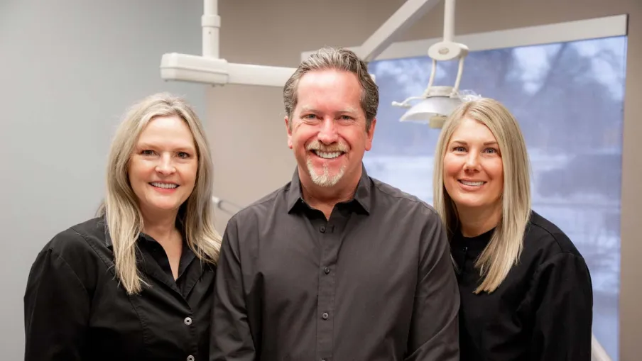 Three smiling dental professionals in black uniforms posing inside a modern dental office with equipment visible.