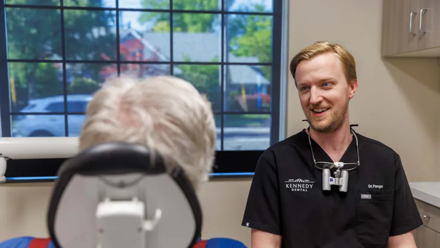 Dentist in black scrubs talking to a patient seated in a dental chair by a window with outdoor view.