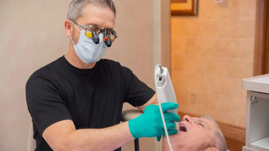 Dentist wearing magnifying glasses and mask using dental scanner on senior male patient's teeth.