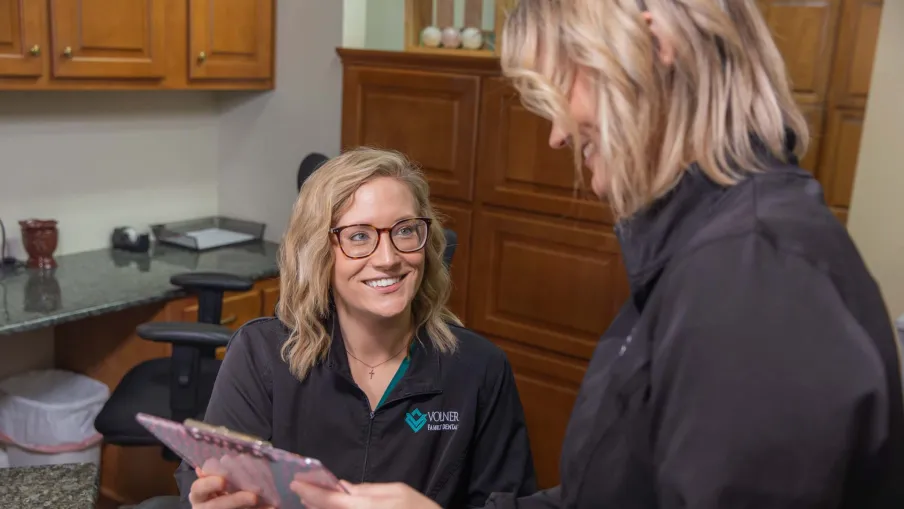 Smiling woman in glasses discusses information on tablet with colleague inside office with wooden cabinets.