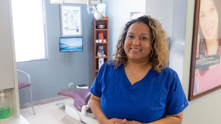 Smiling female dental professional in blue scrubs standing inside a modern dental clinic room.