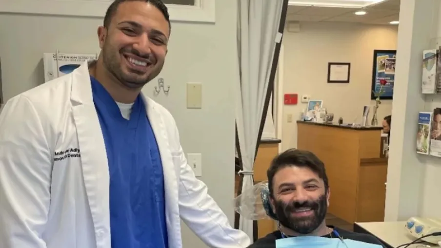 Smiling dentist in blue scrubs and white coat stands next to happy male patient in dental chair.