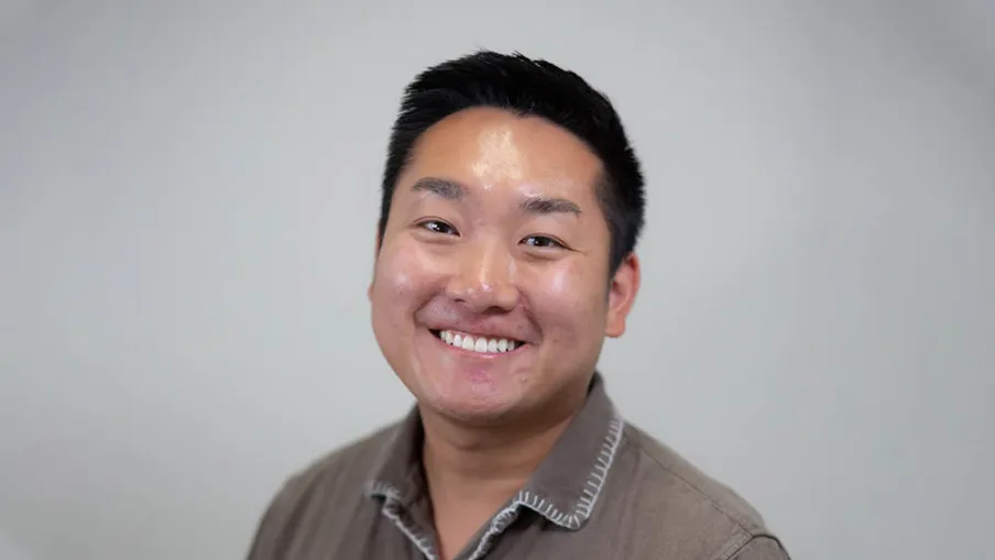 Smiling young Asian man wearing a brown button-up shirt against a plain light gray background.