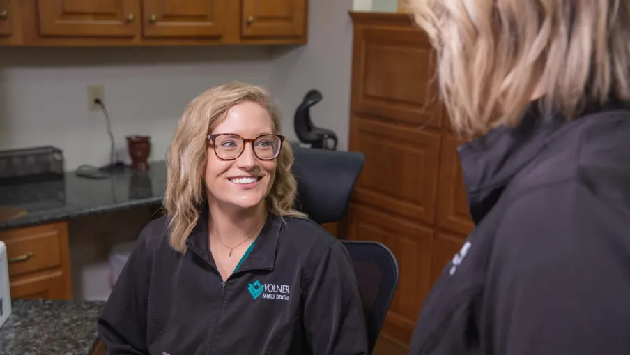 Smiling dental professional wearing glasses and a Volner Family Dental jacket in office setting