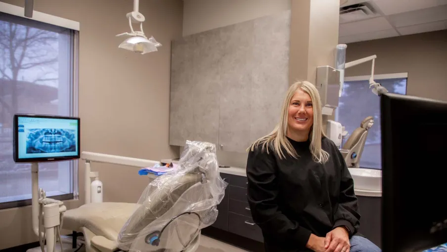 Smiling female dentist in black scrubs sits in a modern dental office with chair and dental X-ray screen.