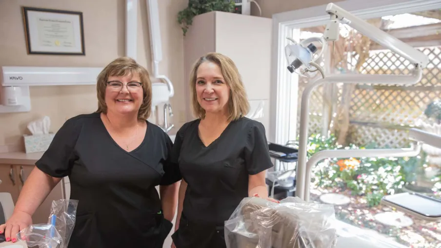 Two female dental hygienists in black scrubs smiling inside a bright dental office with equipment and window view.