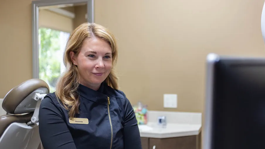 Smiling female dental professional named Danielle sitting in a dental office near patient chair and computer screen